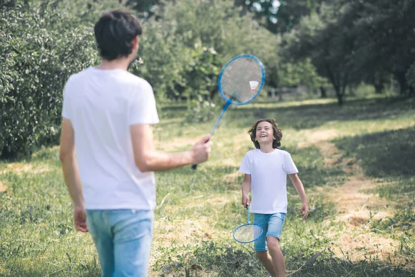 Ciemnowłosy chłopiec i jego ojciec bawią się w badmintona w parku — Zdjęcie stockowe