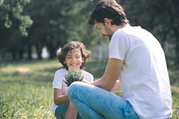 Koyu saçlı çocuk gülümsüyor ve çiçekleri tutuyor, babası onun yanında oturuyor. — Stok fotoğraf