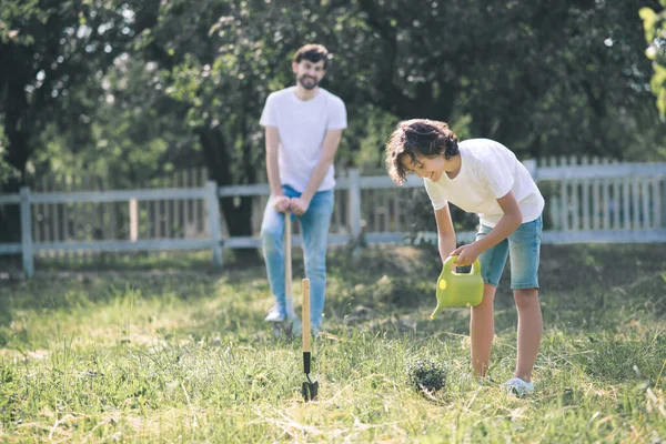 Niño de pelo oscuro regando la planta, su padre de pie cerca — Foto de Stock