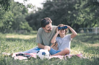 Koyu saçlı çocuk babasının yanında oturmuş dürbün izliyor.