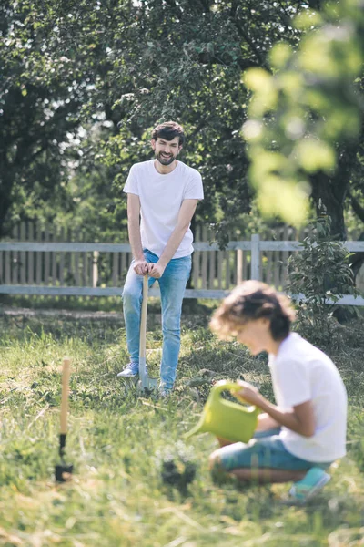 Dark-haired boy watering the plant, his father watching him and smiling — Stock Photo, Image