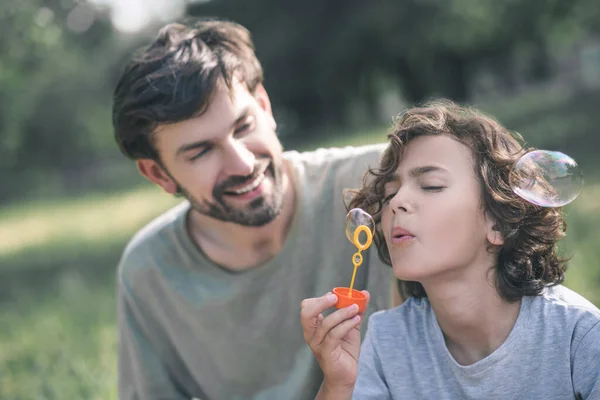 Lindo chico soplando las burbujas, su padre sonriendo — Foto de Stock