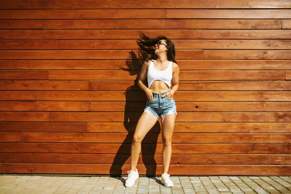 Fit μελαχρινή κοπέλα σε σορτς στέκεται κοντά στον ξύλινο τοίχο — Φωτογραφία Αρχείου