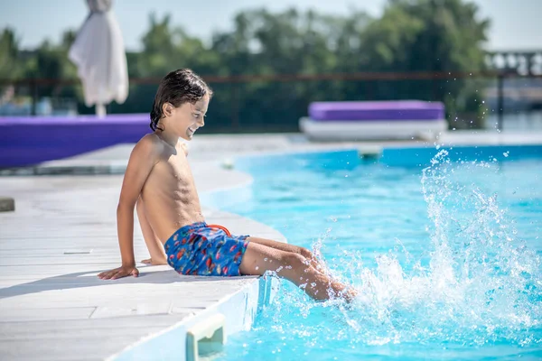 Menino de cabelos escuros sentado na borda da piscina — Fotografia de Stock