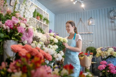 Gülümseyen kahverengi saçlı, at kuyruklu kadın çiçek standında duruyor, gülleri seçiyor.