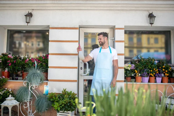 Young bearded male in apron standing at the door of flower shop, holding water spray