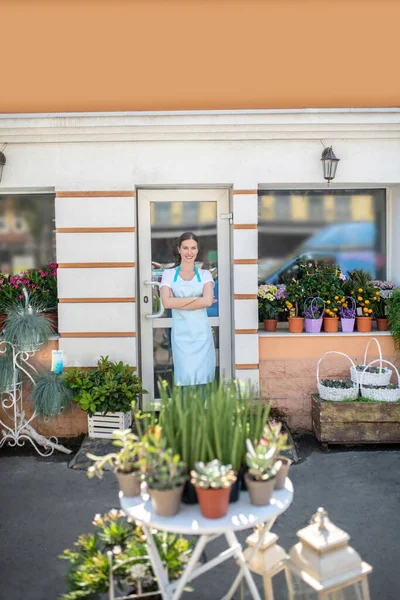 Söt kvinna i förkläde står framför blomsteraffär med armarna korsar — Stockfoto