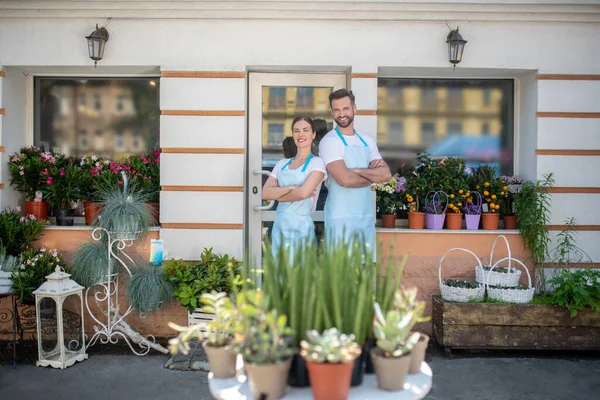 Bearded male and brown-haired female standing proudly outside flower shop, back to back with crossed arms, smiling