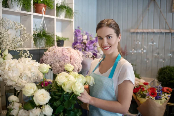 Leende brunhårig hona i förkläde som står framför blomställningen och vidrör rosor — Stockfoto