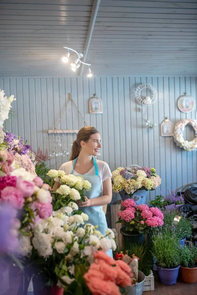 Lächelnde braunhaarige Frau, die am Blumenstand steht, weiße Rosen berührt, seitlich blickt — Stockfoto