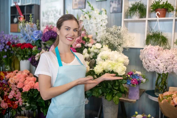 Mujer de cabello castaño en delantal de pie en el puesto de flores, mostrando flores —  Fotos de Stock