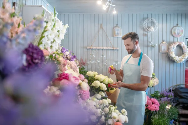Glimlachende bebaarde jonge man in schort sproeien rozen in de voorkant van de bloemenstandaard — Stockfoto
