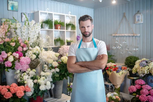 Floristería barbuda en delantal de pie delante de soporte de flores con brazos cruzados —  Fotos de Stock