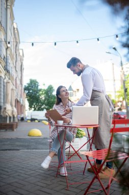 Kahverengi saçlı kadın sakallı garsonla menüyü tartışıyor.