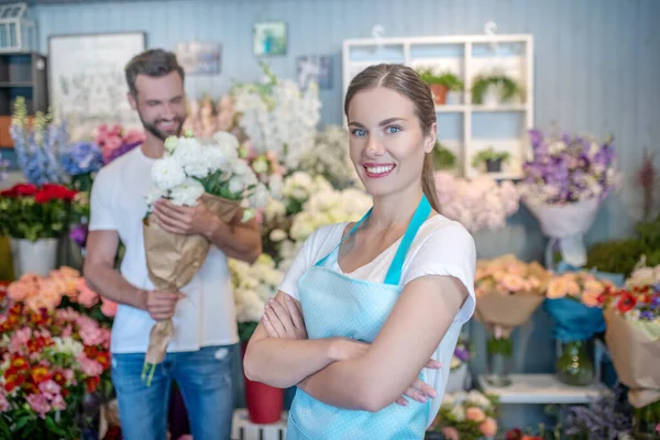 Очаровательная флористка скрещивает руки, мужчина стоит с букетом белых цветов позади нее — стоковое фото