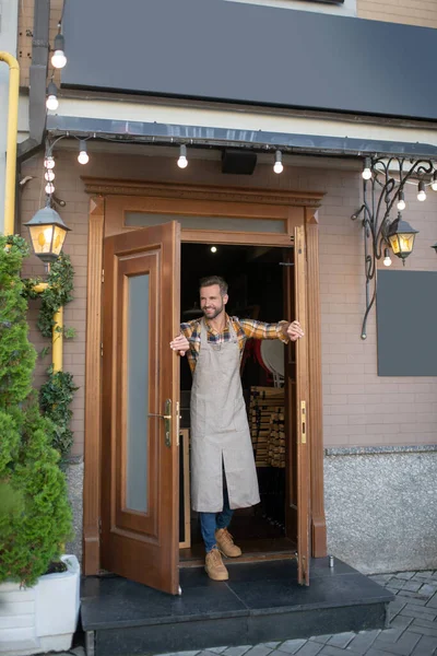 Varón barbudo en delantal gris de pie en la entrada de la cafetería, sosteniendo puertas — Foto de Stock