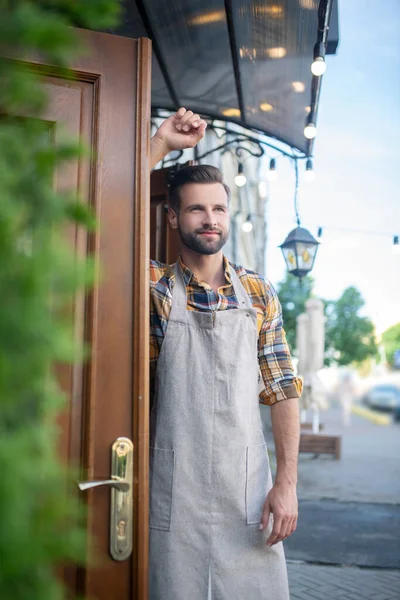 Gri önlüklü sakallı erkek kafe girişinde duruyor, kapıya yaslanıyor. — Stok fotoğraf