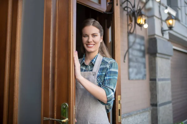 Χαρούμενη σερβιτόρα με γκρι ποδιά που ανοίγει ξύλινη πόρτα. — Φωτογραφία Αρχείου