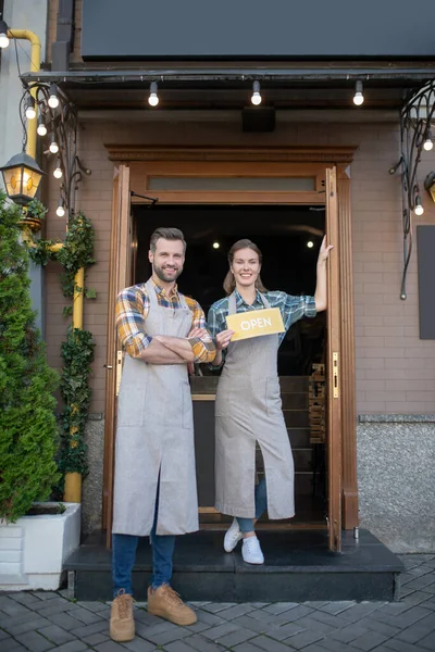 Νεαρή σερβιτόρα και γενειοφόρος σερβιτόρος στέκονται στην πόρτα, κρατώντας ανοιχτή την πινακίδα, χαμογελώντας — Φωτογραφία Αρχείου