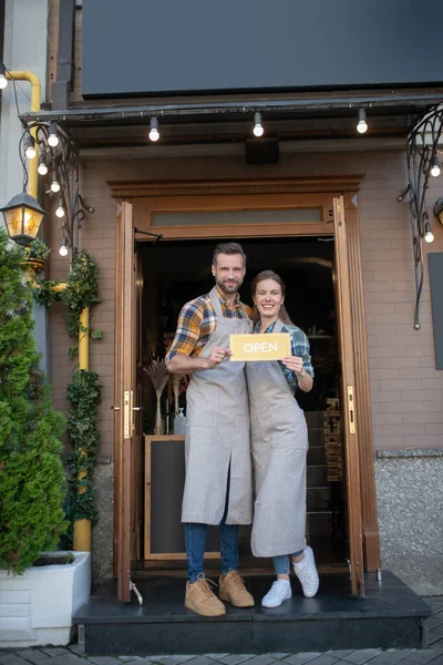 Νεαρή σερβιτόρα και γενειοφόρος σερβιτόρος στέκονται στην είσοδο, κρατώντας κίτρινη ανοιχτή πινακίδα, χαμογελώντας — Φωτογραφία Αρχείου