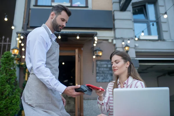 Waiter holding payment machine, brown-haired female paying bill with smartphone