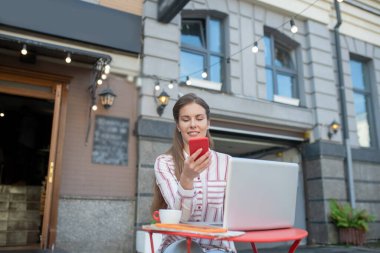 Kahverengi saçlı kadın akıllı telefonu kontrol ediyor, açık hava kafede masada oturuyor.