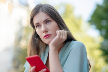 Akıllı telefonunu tutan, çenesini elinde tutan üzgün, tatlı bir kadın.