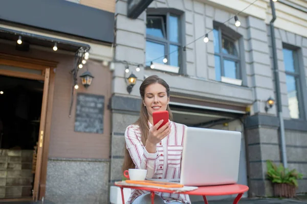 Коричнево-волохата жінка перевіряє смартфон, сидячи за столом у кафе під відкритим небом — стокове фото