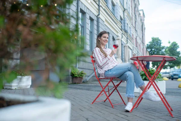 Mujer sonriente sentada con las piernas cruzadas, disfrutando del vino tinto en la cafetería — Foto de Stock