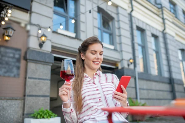 Gülümseyen bir kadın açık hava kafede kırmızı şarap içip telefonuna bakıyor. — Stok fotoğraf