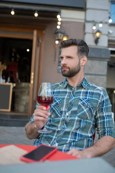 Bearded male sitting in open air cafe, holding glass of red wine — Stock Photo, Image