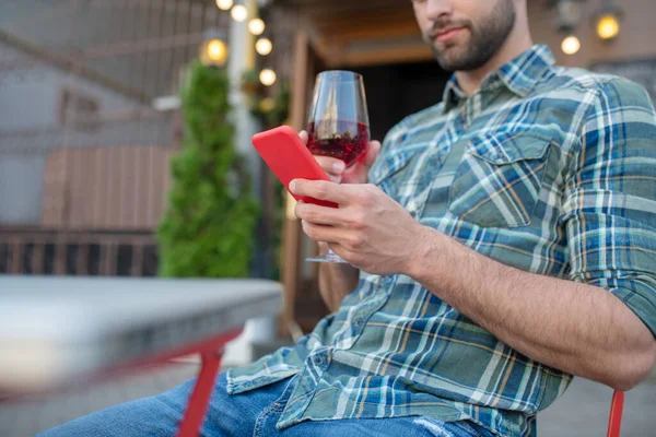 Бородатый красивый мужчина сидит в кафе на открытом воздухе, пьет красное вино, проверяет телефон — стоковое фото