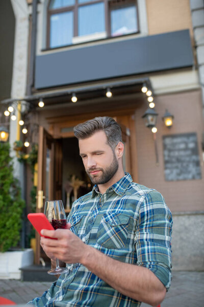 Bearded handsome male sitting in open air cafe, drinking red wine, looking at his phone