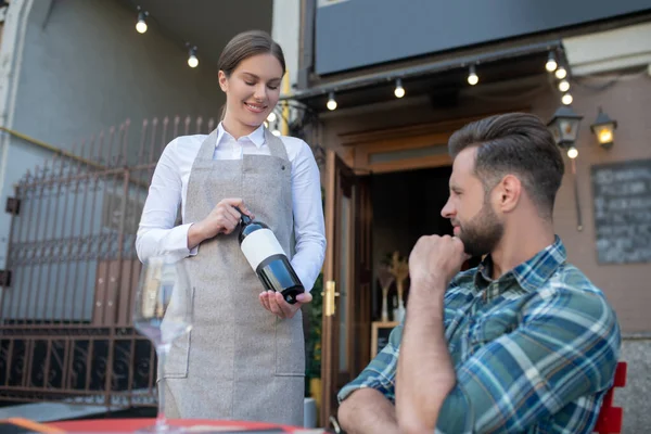 Alegre camarera mostrando botella de vino a hombre barbudo, agujereando su barbilla — Foto de Stock