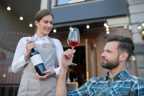 Skäggig hane undersöker vinglas, väljer, ler servitris håller vin flaska — Stockfoto