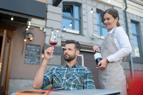 Bearded male looking at wine glass, choosing, smiling waitress holding wine bottle — Stock Photo, Image