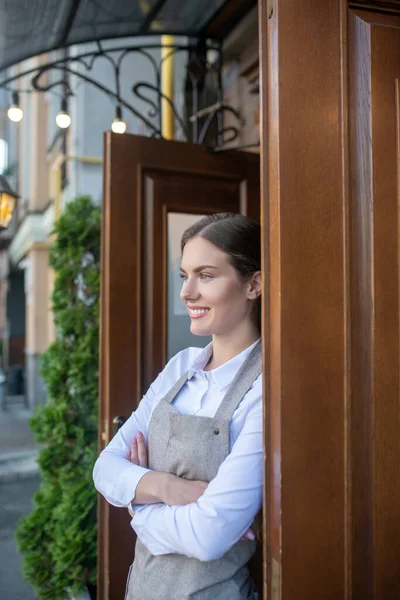 Smiling cute waitress leaning on door with her arms crossed — Stock Photo, Image