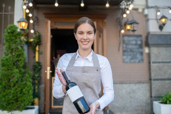 Sorrindo garçonete bonito em avental cinza segurando garrafa de vinho — Fotografia de Stock