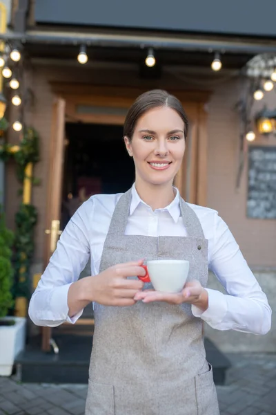 Lachende schattige serveerster in grijs schort met een kopje koffie — Stockfoto