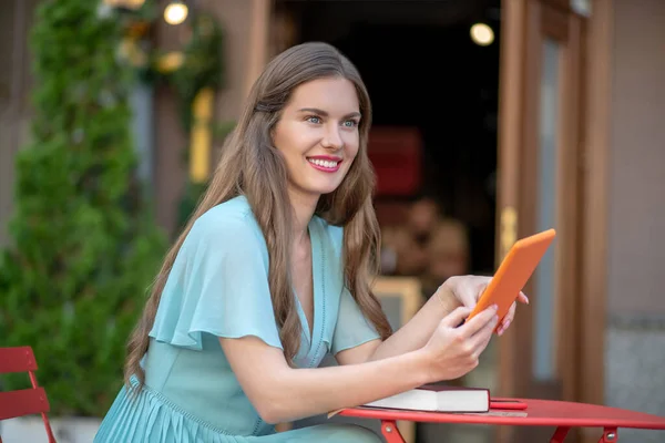 Romantische Frau in blauem Kleid sitzt im Café unter freiem Himmel, hält orangefarbene Tablette in der Hand und lächelt — Stockfoto