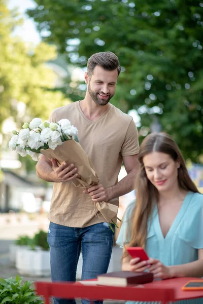 Молода жінка сидить за столом, перевіряючи свій телефон, бородатий чоловік приходить до неї з квітами — стокове фото