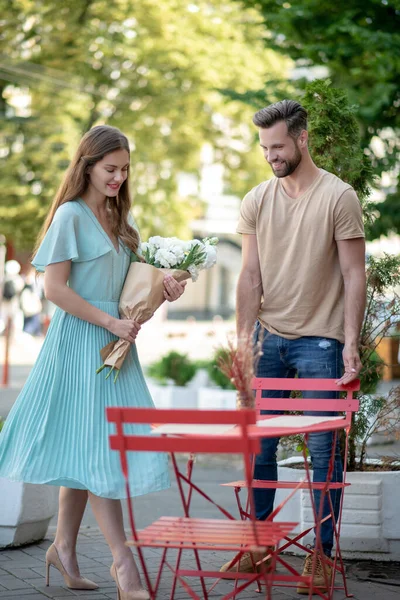 Bearded male pulling red chair for smiling young female with bouquet of white flowers — Stock Photo, Image