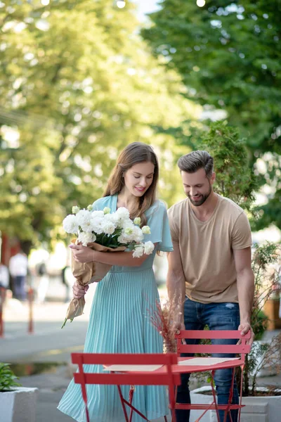 Barbudo masculino puxando cadeira vermelha para feminino com buquê de flores brancas, ajudando-a a sentar-se — Fotografia de Stock