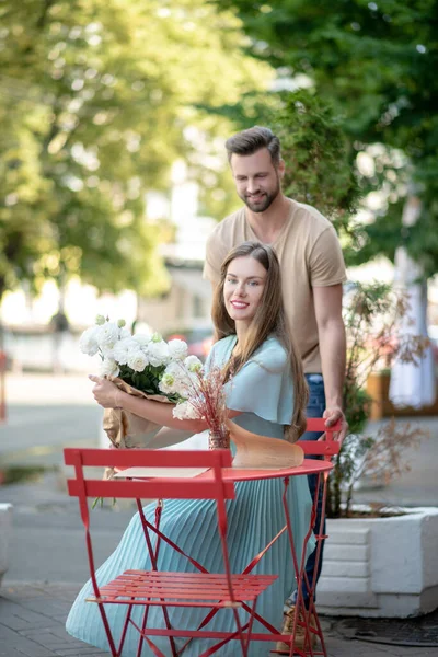 Varón barbudo ayudando a la hembra joven con ramo de flores blancas a sentarse a la mesa —  Fotos de Stock