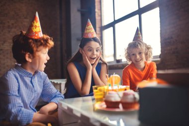 Üç çocuk pastaya bakıyor ve heyecanlı görünüyorlar.