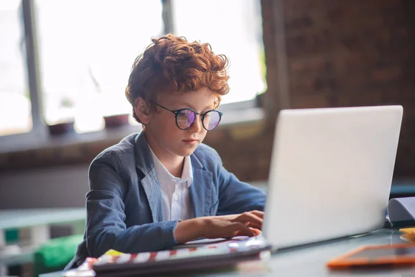 Rudovlasý chlapec sedí u laptopu a vypadá zaneprázdněně — Stock fotografie