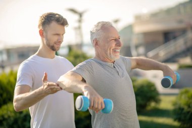 Elinde dambıl tutan gülümseyen olgun bir adam, ona yardım eden sakallı bir eğitmen.