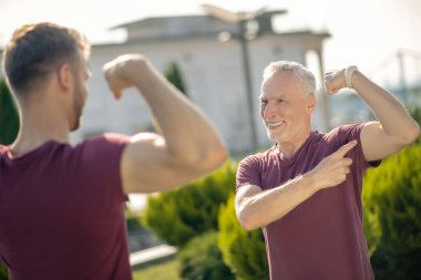 Genç erkek ve gri saçlı adam ayakta, kaslarını gösteriyor, gülümsüyor.