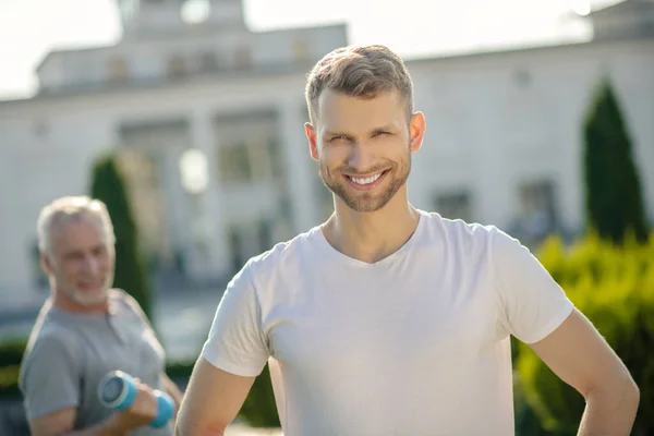 Joven barbudo macho sonriente, de pelo gris macho sosteniendo mancuerna detrás —  Fotos de Stock