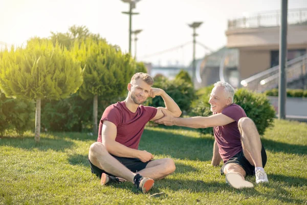 Gråhårig man rör skäggiga manliga biceps, båda sitter på gräs — Stockfoto